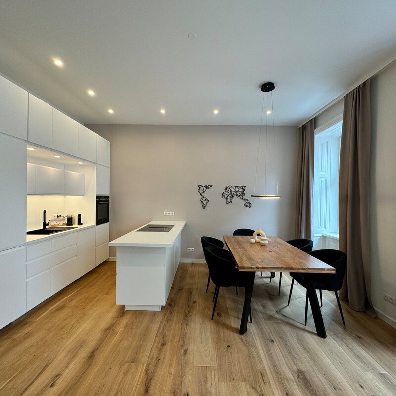 The open living and dining area features modern furnishings, a fully equipped kitchen, a large dining table, and stylish furniture. The large windows allow plenty of natural light, creating a bright and inviting atmosphere.