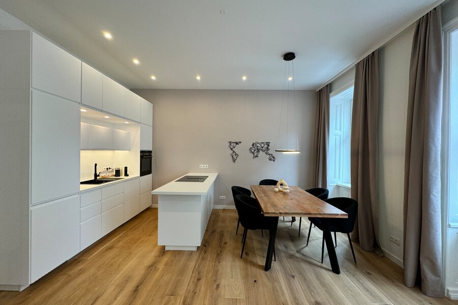 The open living and dining area features modern furnishings, a fully equipped kitchen, a large dining table, and stylish furniture. The large windows allow plenty of natural light, creating a bright and inviting atmosphere.