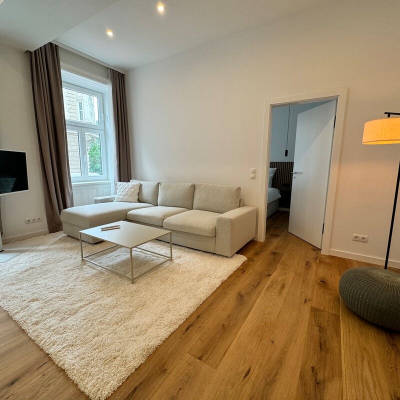 The living room features a comfortable pull-out couch, a modern flat-screen TV, and stylish lighting. The warm wooden floors and large windows create an inviting and cozy atmosphere.