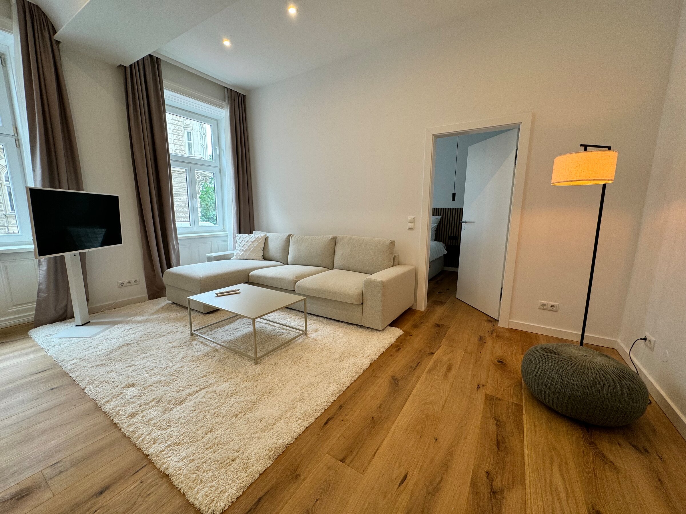 The living room features a comfortable pull-out couch, a modern flat-screen TV, and stylish lighting. The warm wooden floors and large windows create an inviting and cozy atmosphere.
