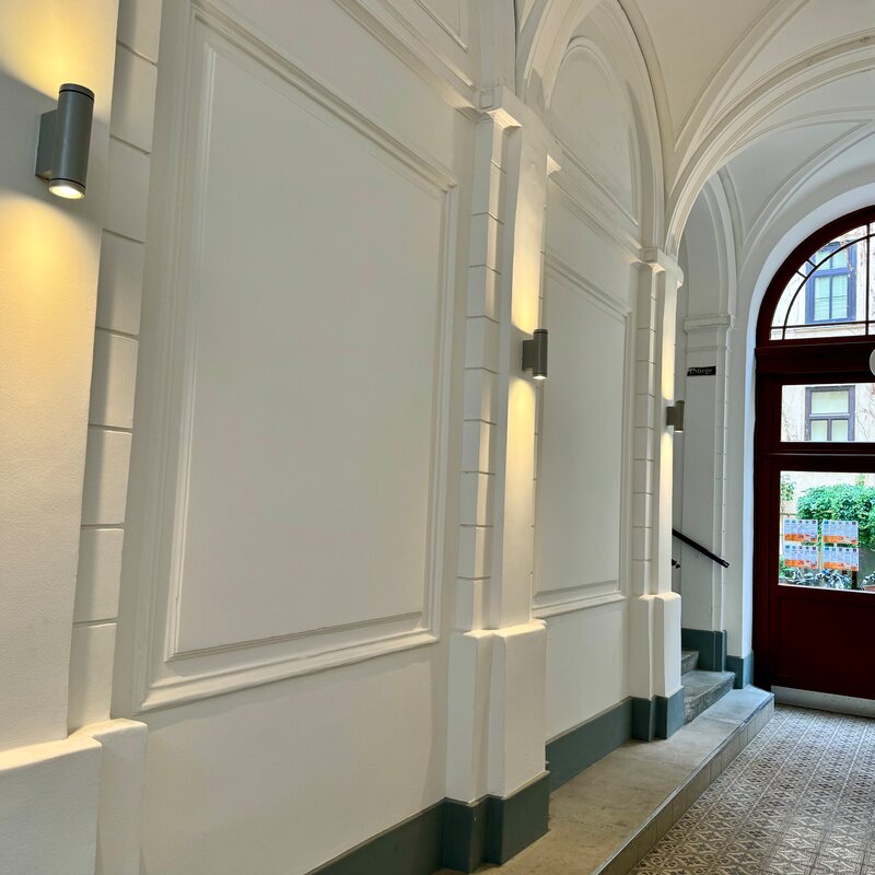 Der eindrucksvolle Hauseingangsbereich führt zur Wohnung und besticht durch seine klassische Architektur und elegante Beleuchtung. Die hohen Decken und die stilvollen Details schaffen ein einladendes Ambiente. Durch die Tür gelangt man in den Hofbereich, wo sich Fahrradstellplätze und Mülltonnen befinden.