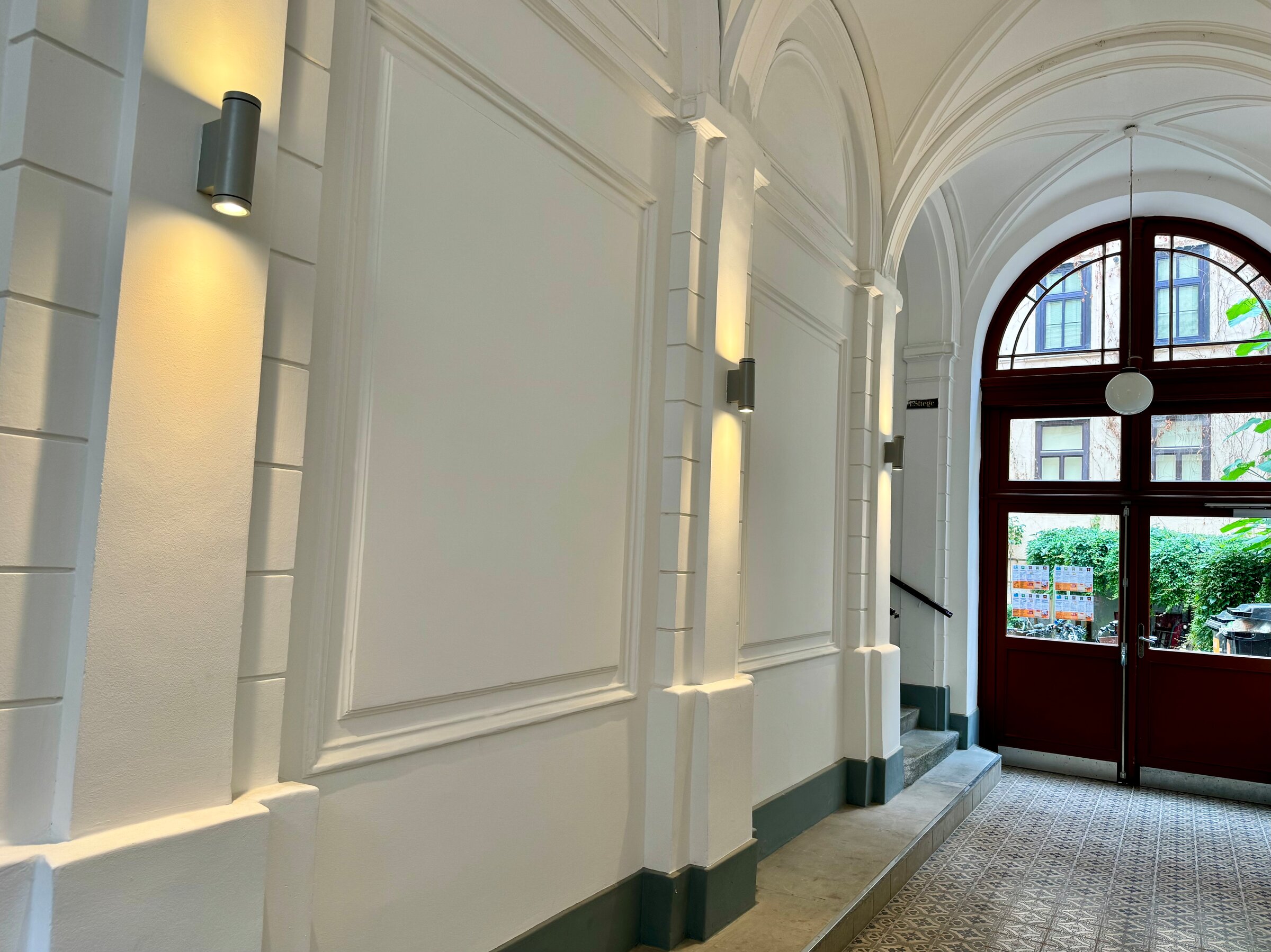 Der eindrucksvolle Hauseingangsbereich führt zur Wohnung und besticht durch seine klassische Architektur und elegante Beleuchtung. Die hohen Decken und die stilvollen Details schaffen ein einladendes Ambiente. Durch die Tür gelangt man in den Hofbereich, wo sich Fahrradstellplätze und Mülltonnen befinden.