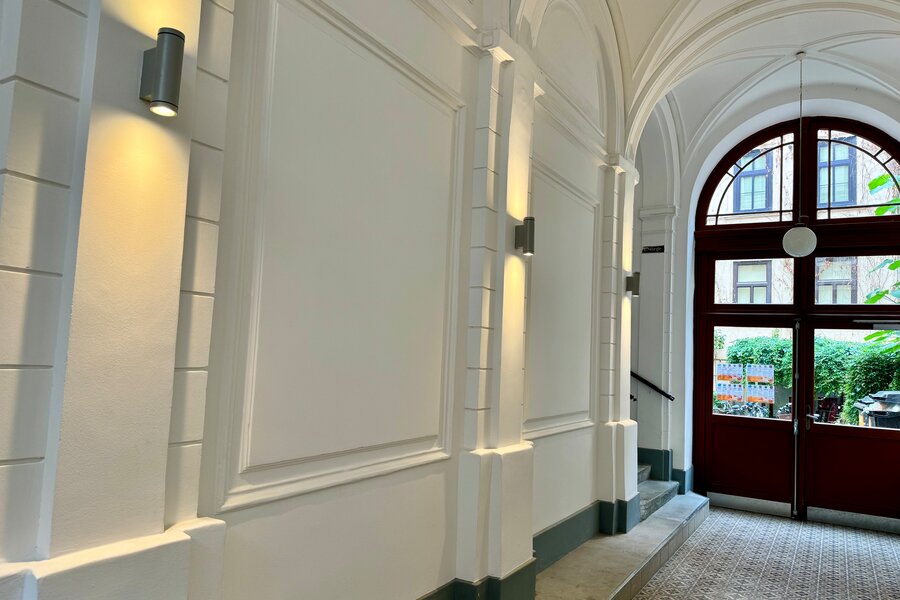 Der eindrucksvolle Hauseingangsbereich führt zur Wohnung und besticht durch seine klassische Architektur und elegante Beleuchtung. Die hohen Decken und die stilvollen Details schaffen ein einladendes Ambiente. Durch die Tür gelangt man in den Hofbereich, wo sich Fahrradstellplätze und Mülltonnen befinden.