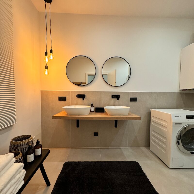 The bathroom is modern and stylishly furnished, featuring two sinks, a spacious shower, and a toilet. A washing machine adds extra convenience. The tasteful lighting and elegant details create a relaxing atmosphere.