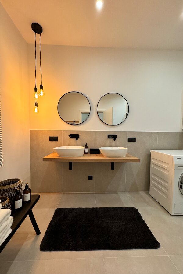 The bathroom is modern and stylishly furnished, featuring two sinks, a spacious shower, and a toilet. A washing machine adds extra convenience. The tasteful lighting and elegant details create a relaxing atmosphere.