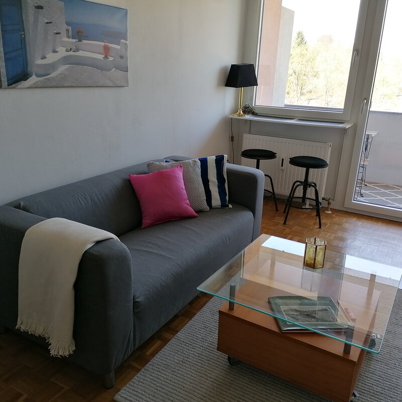 Wohnraum, ausgestattet mit einer Couch, einem Tisch, Theke am Fenster mit 2 Hockern, Schubladkasten, großer Kleiderschrank und einer Schlafnische mit einem Bett 140x200