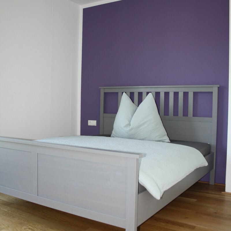 Helles Schlafzimmer mit grauem Holzbett & lila Akzentwand – 4-Zimmer-Wohnung, Kurzzeitmiete Innsbruck, Wohnen auf Zeit 1–6 Monate