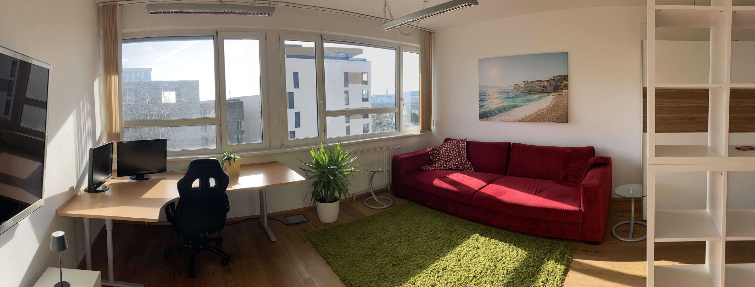 Helles Wohnzimmer mit Arbeitsbereich, rotem Sofa & Grünpflanze – 1-Zimmer Wohnung, Kurzzeitmiete Linz, Wohnen auf Zeit 1 bis 6 Monate.