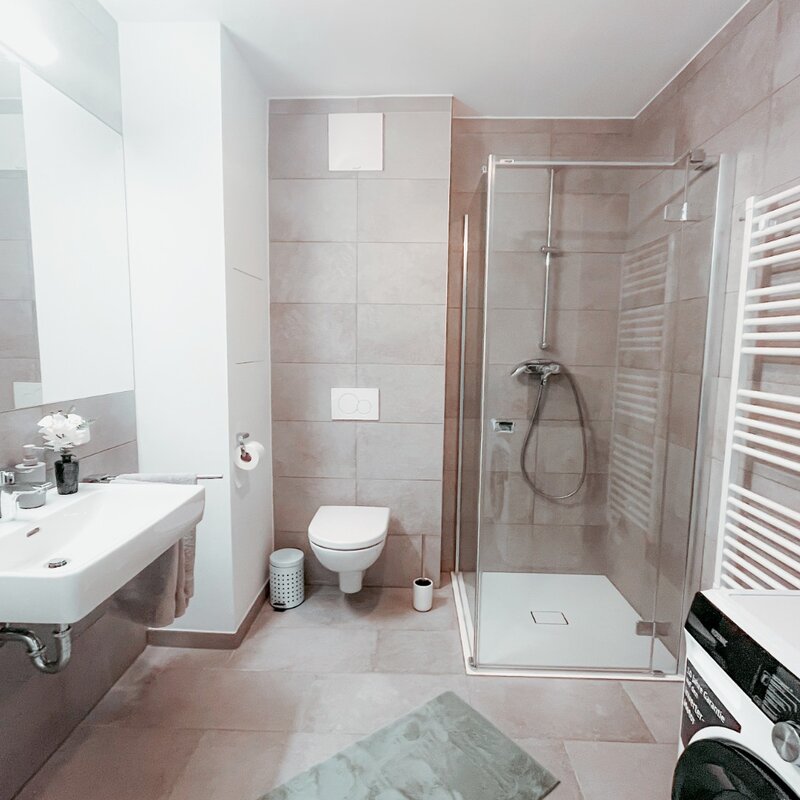 Badezimmer mit großem Spiegel, Waschbecken, Duschkabine, Waschmaschine mit Wasch- und Trockenfunktion, eingerichtet im eleganten Stil. | © All rights reserved