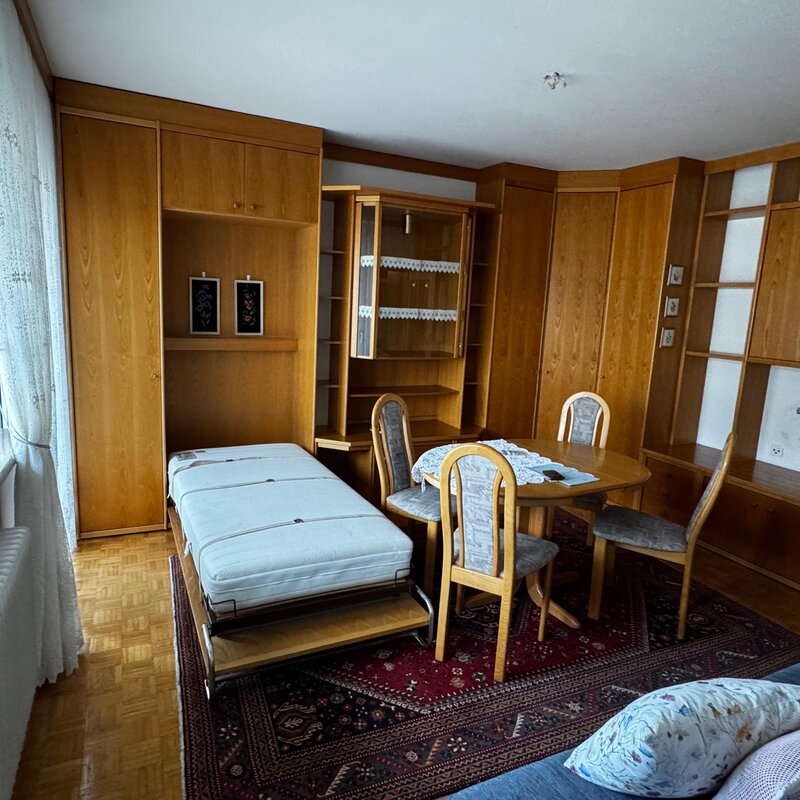 Der Wohn-/Schlafraum ist mit Einbaumöbeln aus Holz ausgestattet und bietet viel Stauraum. Das ausklappbare Bett sorgt für eine flexible Nutzung des Raums. Ein großer Esstisch mit Stühlen macht den Bereich auch als Arbeits- oder Essplatz nutzbar. Helles Fenster mit Gardinen und Parkettboden sorgen für eine angenehme Atmosphäre. | © Stefan Gabriel