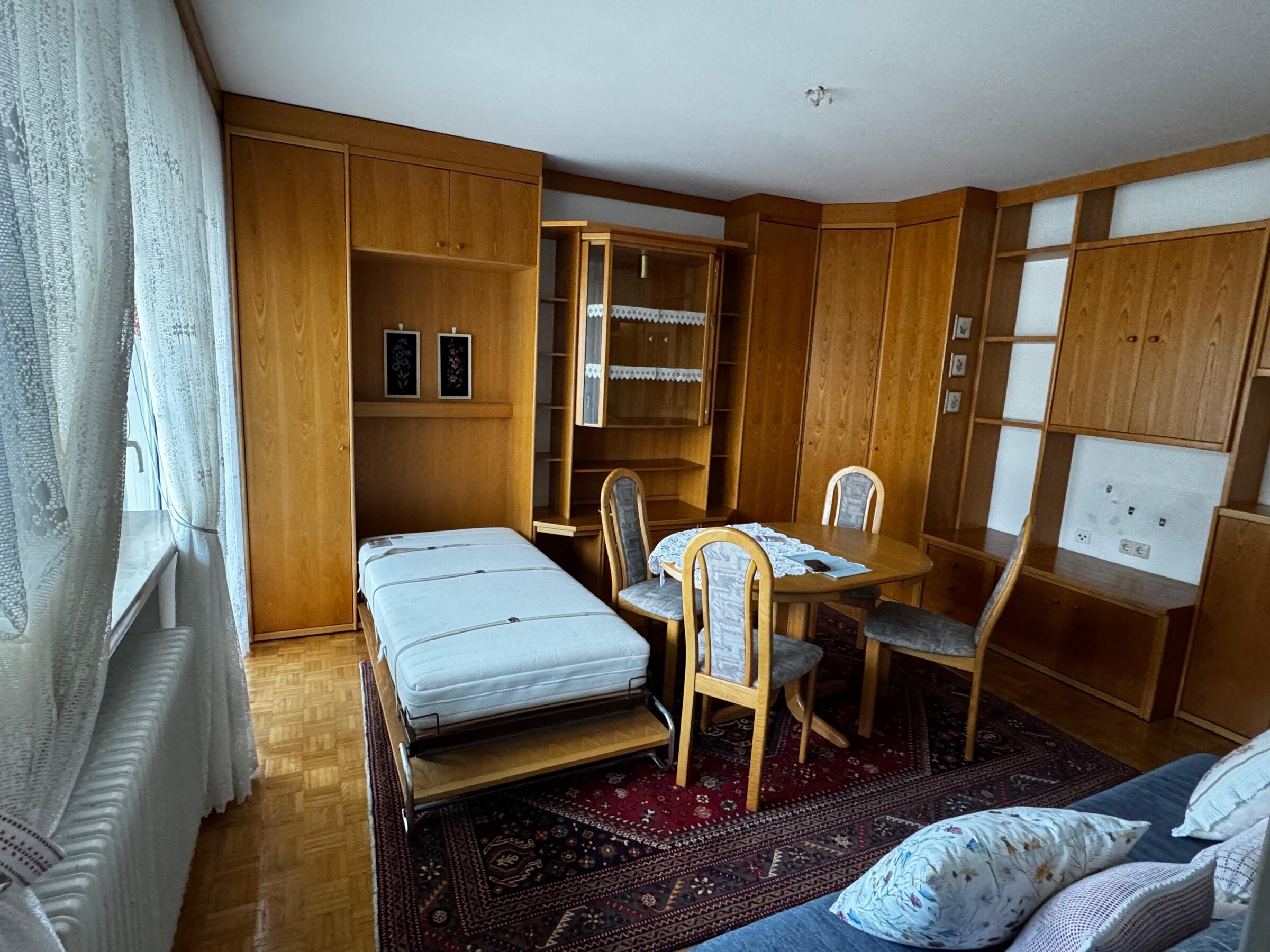 Der Wohn-/Schlafraum ist mit Einbaumöbeln aus Holz ausgestattet und bietet viel Stauraum. Das ausklappbare Bett sorgt für eine flexible Nutzung des Raums. Ein großer Esstisch mit Stühlen macht den Bereich auch als Arbeits- oder Essplatz nutzbar. Helles Fenster mit Gardinen und Parkettboden sorgen für eine angenehme Atmosphäre. | © Stefan Gabriel