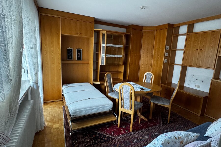 Der Wohn-/Schlafraum ist mit Einbaumöbeln aus Holz ausgestattet und bietet viel Stauraum. Das ausklappbare Bett sorgt für eine flexible Nutzung des Raums. Ein großer Esstisch mit Stühlen macht den Bereich auch als Arbeits- oder Essplatz nutzbar. Helles Fenster mit Gardinen und Parkettboden sorgen für eine angenehme Atmosphäre. | © Stefan Gabriel