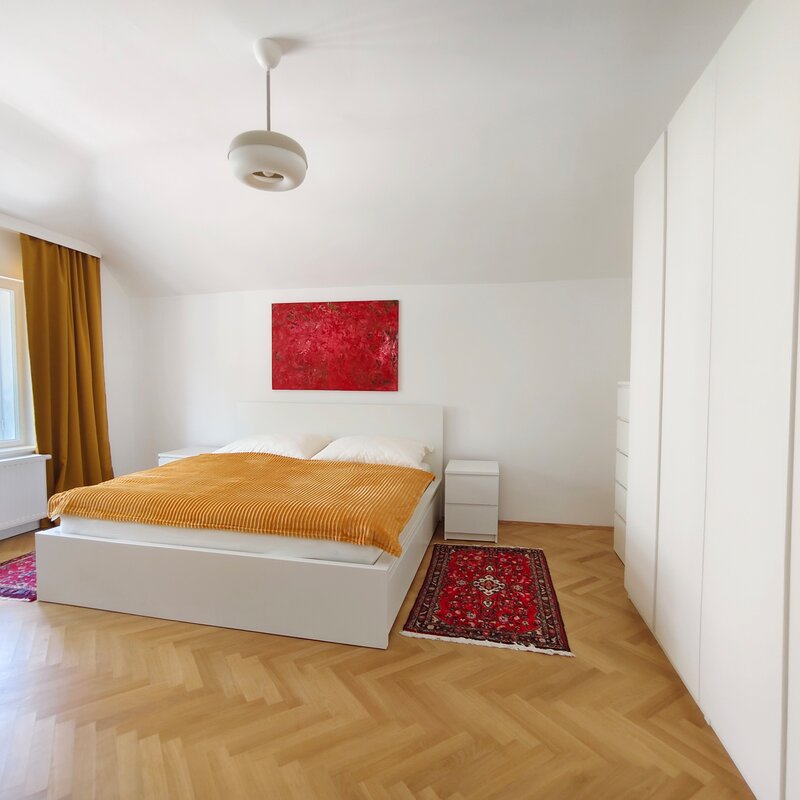 The double bed is between a big window and a dresser beside wardrobes, the herringbone parquet flooring is partly covered with carpets. 