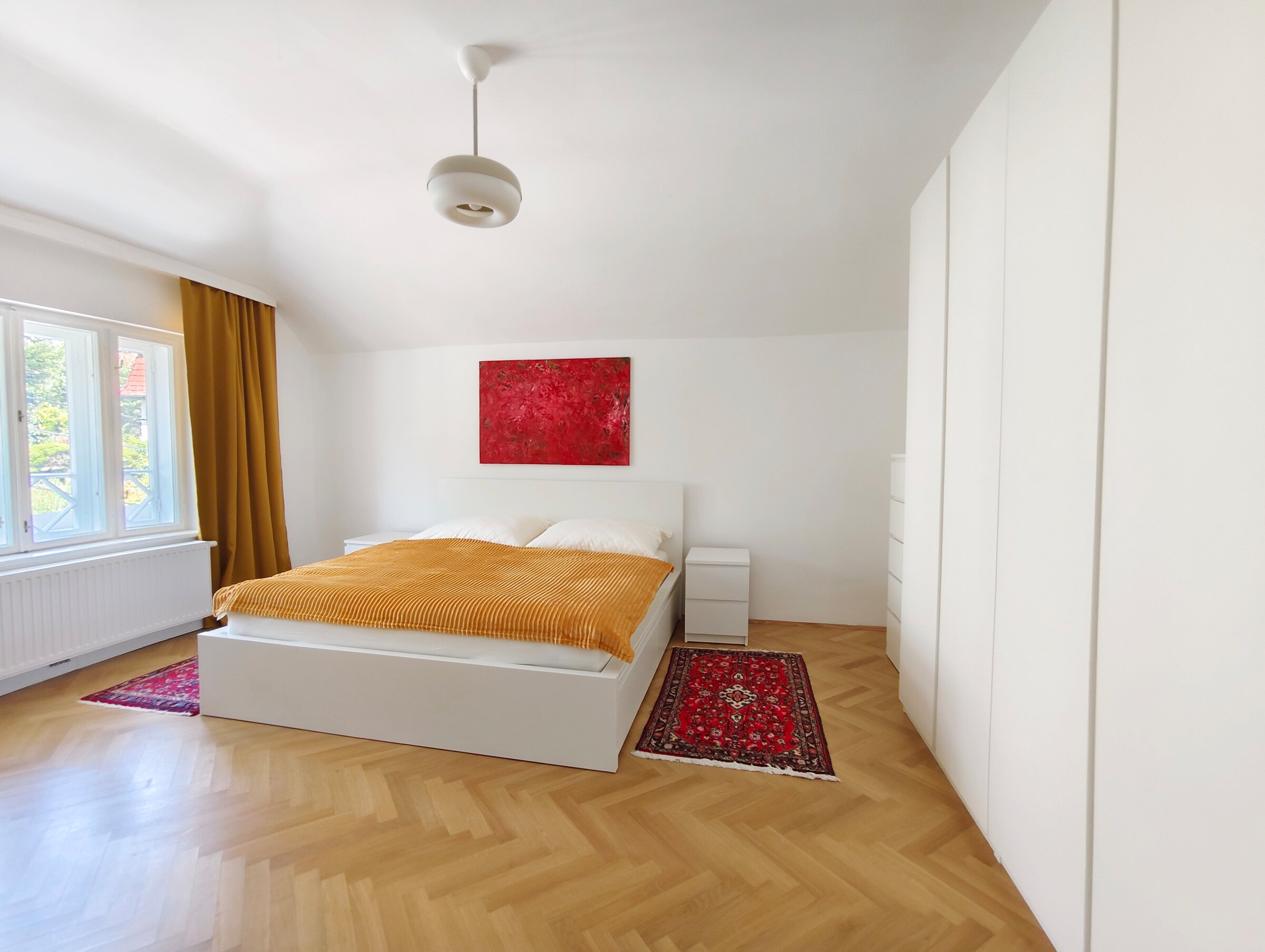 The double bed is between a big window and a dresser beside wardrobes, the herringbone parquet flooring is partly covered with carpets. 