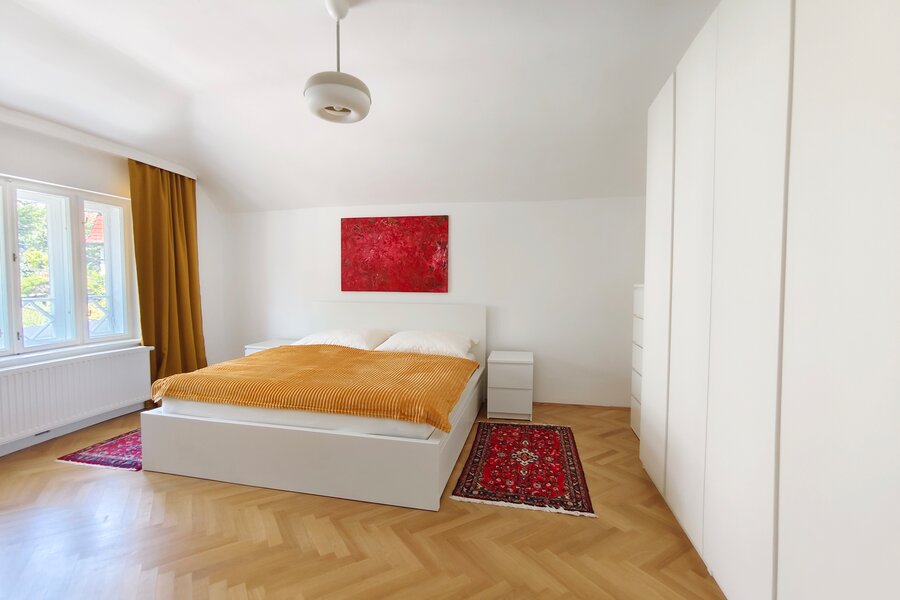 The double bed is between a big window and a dresser beside wardrobes, the herringbone parquet flooring is partly covered with carpets. 