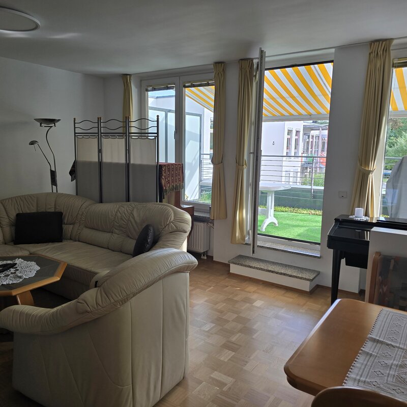 Helles Wohnzimmer mit Ecksofa und gemütlicher Sitzgruppe. Große Fenster mit gelb-weiß gestreiften Markisen bieten Zugang zur begrünten Terrasse. Ein kleines Klavier steht an der Wand, daneben eine schlichte Abtrennung zur Essecke.