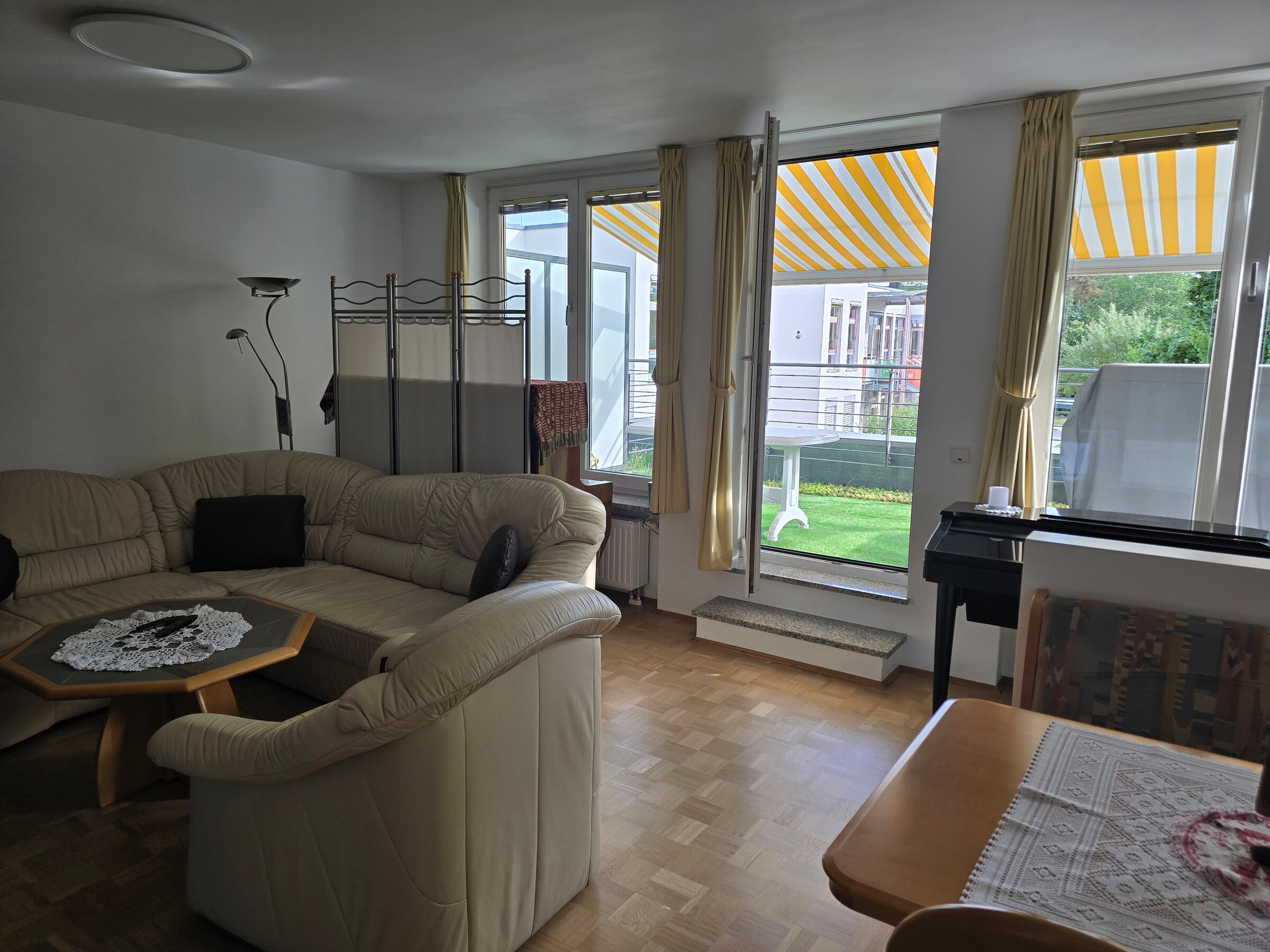 Helles Wohnzimmer mit Ecksofa und gemütlicher Sitzgruppe. Große Fenster mit gelb-weiß gestreiften Markisen bieten Zugang zur begrünten Terrasse. Ein kleines Klavier steht an der Wand, daneben eine schlichte Abtrennung zur Essecke.