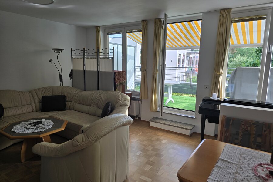 Helles Wohnzimmer mit Ecksofa und gemütlicher Sitzgruppe. Große Fenster mit gelb-weiß gestreiften Markisen bieten Zugang zur begrünten Terrasse. Ein kleines Klavier steht an der Wand, daneben eine schlichte Abtrennung zur Essecke.