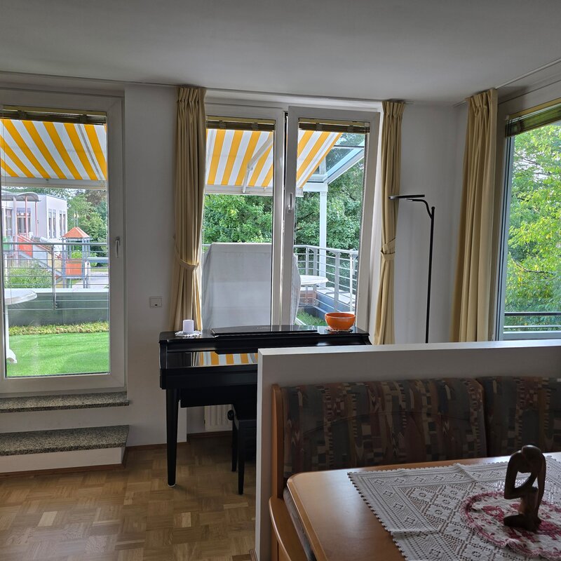 Der kombinierte Wohn- und Essbereich bietet drei große Öffnungen mit Blick ins Grüne – zwei Fenster und eine Terrassentür zur südlichen Terrasse. Ganz rechts führt eine Tür direkt zur überdachten Loggia. Helle Vorhänge, Parkettboden und viel Tageslicht sorgen für eine freundliche Atmosphäre.