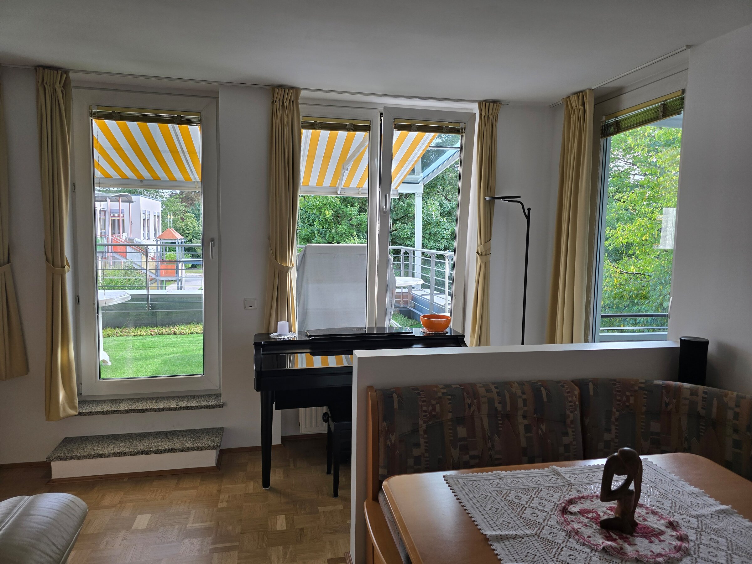 Der kombinierte Wohn- und Essbereich bietet drei große Öffnungen mit Blick ins Grüne – zwei Fenster und eine Terrassentür zur südlichen Terrasse. Ganz rechts führt eine Tür direkt zur überdachten Loggia. Helle Vorhänge, Parkettboden und viel Tageslicht sorgen für eine freundliche Atmosphäre.