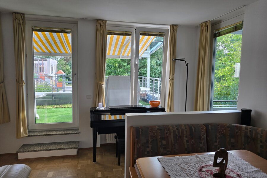 Der kombinierte Wohn- und Essbereich bietet drei große Öffnungen mit Blick ins Grüne – zwei Fenster und eine Terrassentür zur südlichen Terrasse. Ganz rechts führt eine Tür direkt zur überdachten Loggia. Helle Vorhänge, Parkettboden und viel Tageslicht sorgen für eine freundliche Atmosphäre.