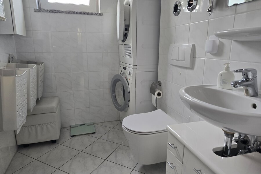 Dieses praktische zweite Badezimmer ist mit einem WC, Waschbecken und einem Waschmaschinen-Trockner-Turm ausgestattet. Ideal für Familien oder Wohngemeinschaften - hier lassen sich Wäschepflege und tägliche Nutzung bequem kombinieren. Helle Fliesen, Tageslichtfenster und durchdachte Stauraumlösungen sorgen für Funktionalität und Komfort.
