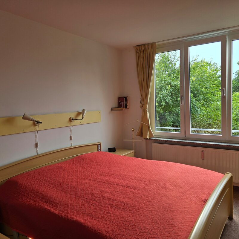 Das freundliche Schlafzimmer ist mit einem großen Fenster zur Westseite ausgestattet und bietet einen schönen, unverbauten Blick ins Grüne. Die Raumgestaltung mit Doppelbett, Nachttischen und Leselampen sorgt für eine gemütliche Atmosphäre.