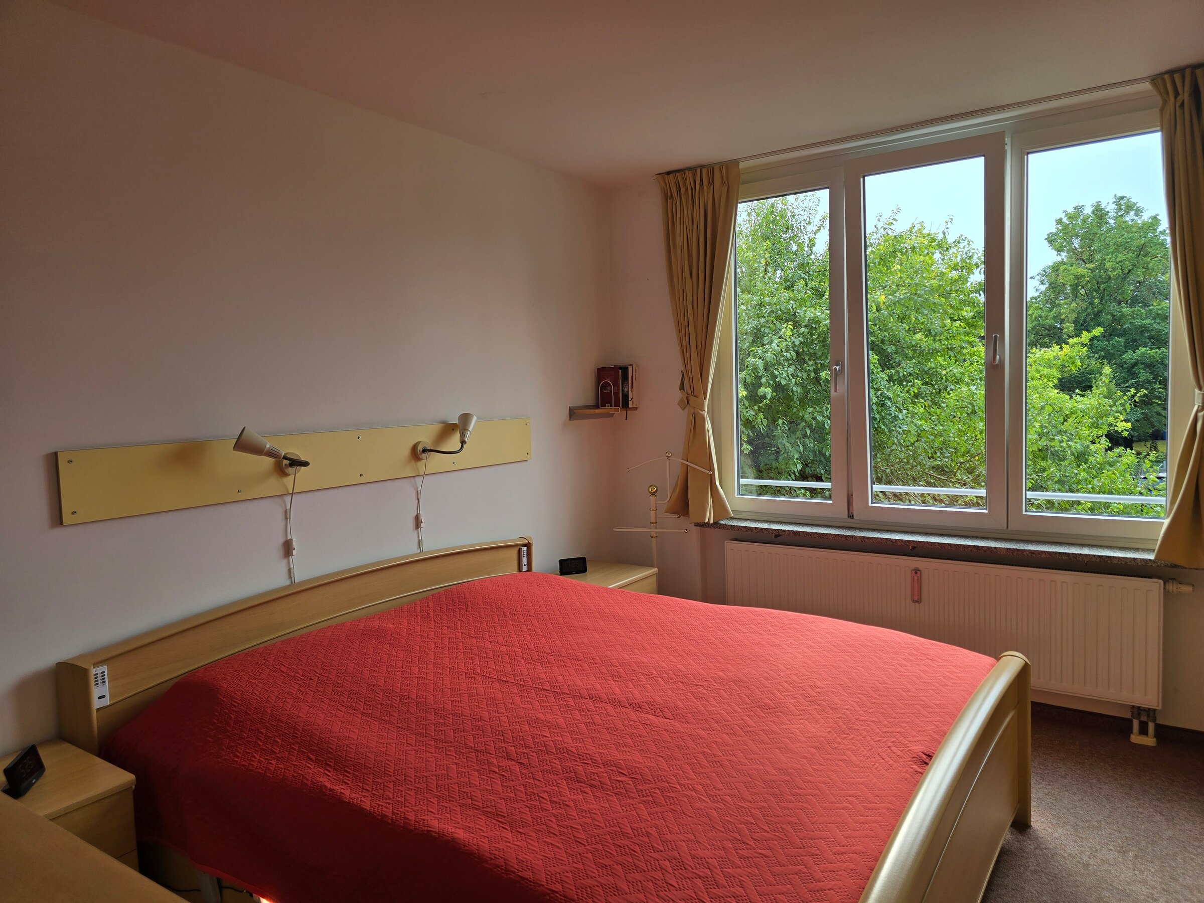 Das freundliche Schlafzimmer ist mit einem großen Fenster zur Westseite ausgestattet und bietet einen schönen, unverbauten Blick ins Grüne. Die Raumgestaltung mit Doppelbett, Nachttischen und Leselampen sorgt für eine gemütliche Atmosphäre.