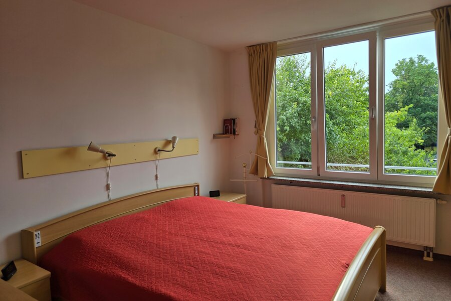 Das freundliche Schlafzimmer ist mit einem großen Fenster zur Westseite ausgestattet und bietet einen schönen, unverbauten Blick ins Grüne. Die Raumgestaltung mit Doppelbett, Nachttischen und Leselampen sorgt für eine gemütliche Atmosphäre.