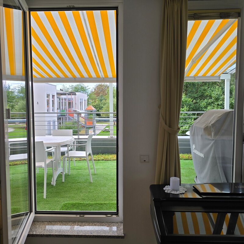 Dieses Bild zeigt die beiden großen Fensteröffnungen im Wohnbereich mit direktem Zugang zur großzügigen, begrünten Terrasse. Im Hintergrund ist der Kindergarten im Erdgeschoss erkennbar. Die Markise bietet Sonnenschutz und unterstreicht die wohnliche Atmosphäre.