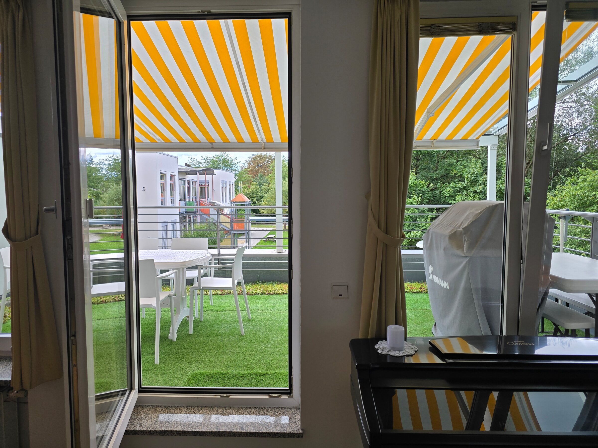 Dieses Bild zeigt die beiden großen Fensteröffnungen im Wohnbereich mit direktem Zugang zur großzügigen, begrünten Terrasse. Im Hintergrund ist der Kindergarten im Erdgeschoss erkennbar. Die Markise bietet Sonnenschutz und unterstreicht die wohnliche Atmosphäre.