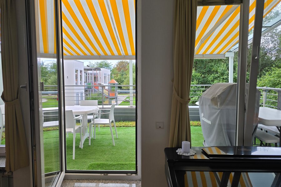 Dieses Bild zeigt die beiden großen Fensteröffnungen im Wohnbereich mit direktem Zugang zur großzügigen, begrünten Terrasse. Im Hintergrund ist der Kindergarten im Erdgeschoss erkennbar. Die Markise bietet Sonnenschutz und unterstreicht die wohnliche Atmosphäre.