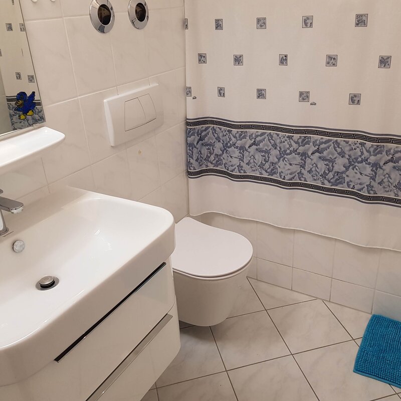 Das Badezimmer ist hell gefliest und mit einem modernen Waschbecken, einem Hänge-WC sowie einem Spiegel ausgestattet. Ein Vorhang mit dezentem Muster und eine farblich passende Badematte sorgen für eine wohnliche Atmosphäre.