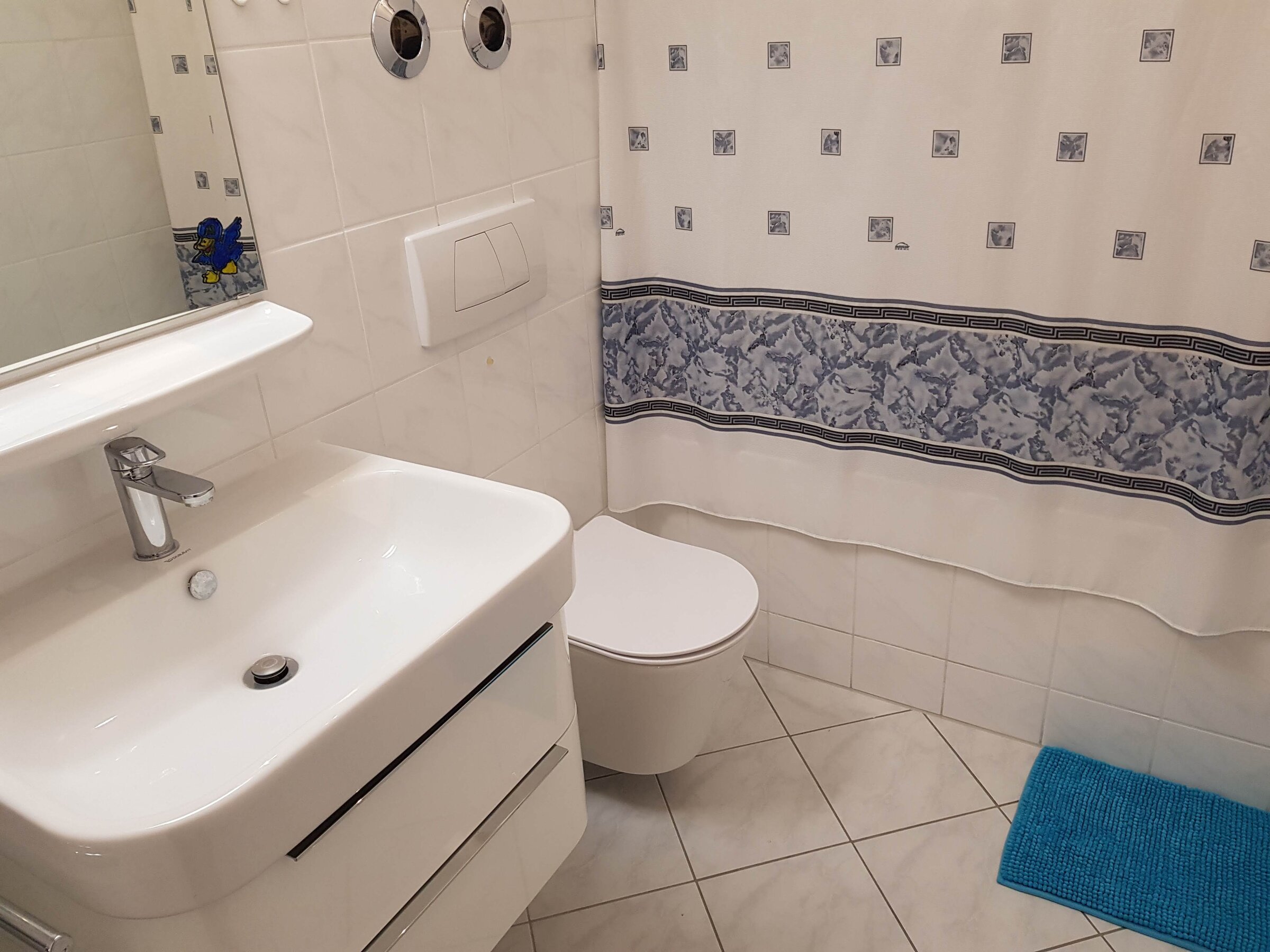 Das Badezimmer ist hell gefliest und mit einem modernen Waschbecken, einem Hänge-WC sowie einem Spiegel ausgestattet. Ein Vorhang mit dezentem Muster und eine farblich passende Badematte sorgen für eine wohnliche Atmosphäre.