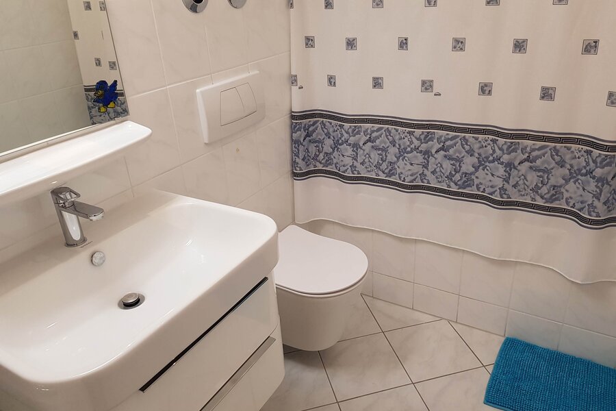 Das Badezimmer ist hell gefliest und mit einem modernen Waschbecken, einem Hänge-WC sowie einem Spiegel ausgestattet. Ein Vorhang mit dezentem Muster und eine farblich passende Badematte sorgen für eine wohnliche Atmosphäre.