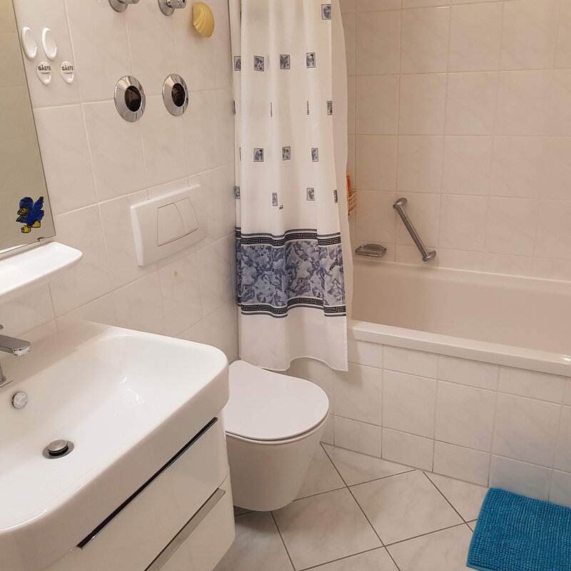 Das Badezimmer verfügt zusätzlich über eine Badewanne mit Haltegriff, ideal für entspannende Momente. Auch hier sorgen helle Fliesen und funktionale Elemente für Komfort und Sauberkeit.