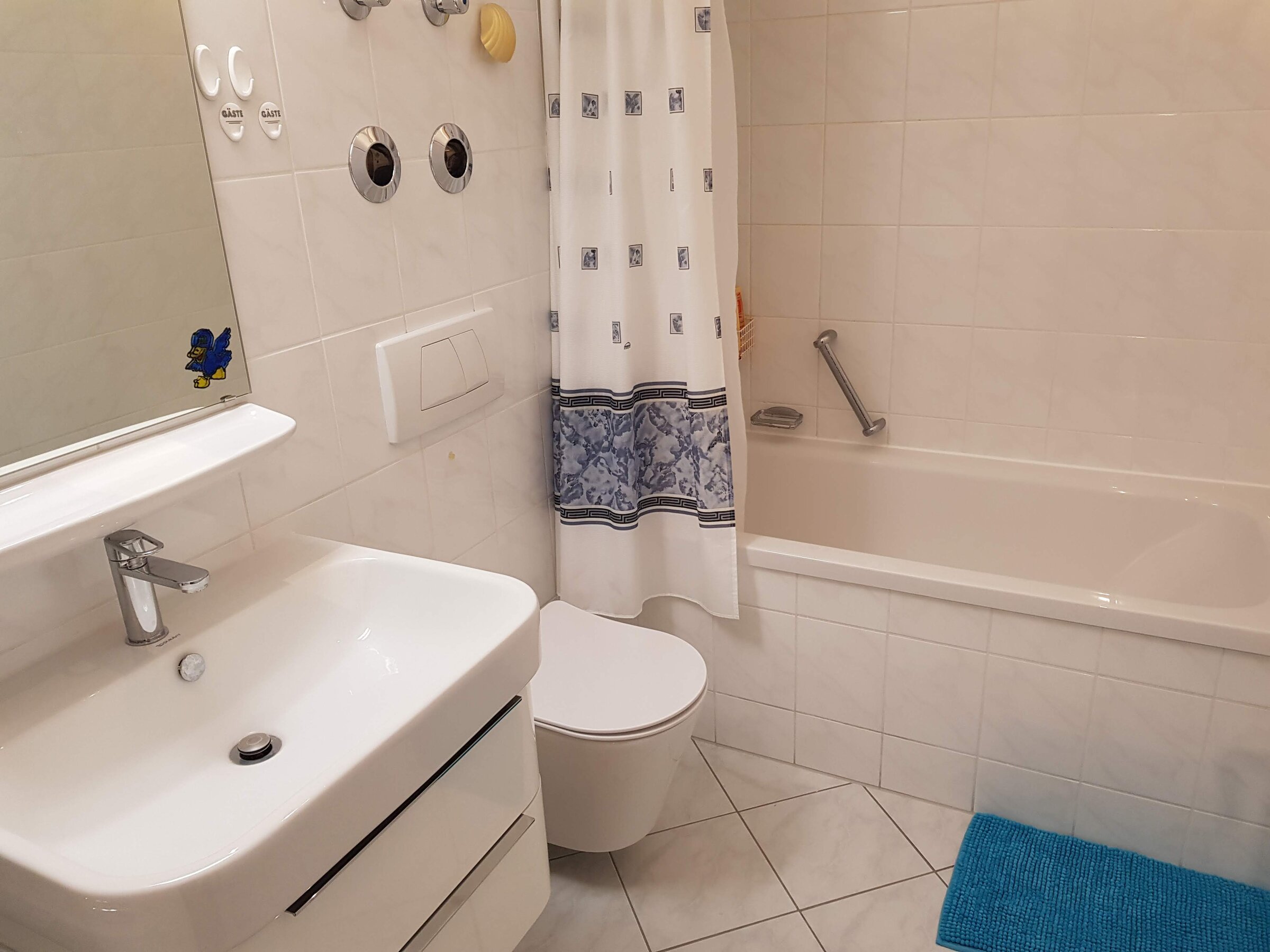 Das Badezimmer verfügt zusätzlich über eine Badewanne mit Haltegriff, ideal für entspannende Momente. Auch hier sorgen helle Fliesen und funktionale Elemente für Komfort und Sauberkeit.