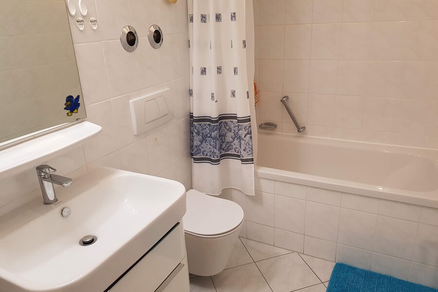 Das Badezimmer verfügt zusätzlich über eine Badewanne mit Haltegriff, ideal für entspannende Momente. Auch hier sorgen helle Fliesen und funktionale Elemente für Komfort und Sauberkeit.