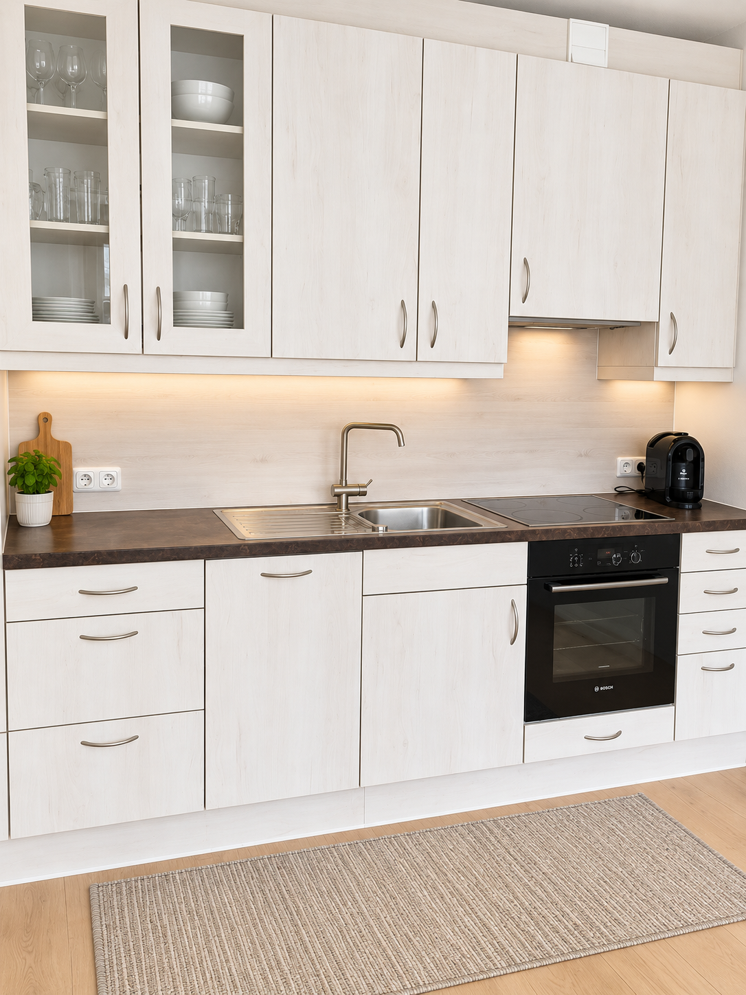 Die helle Küchenzeile bietet alles, was man für den Alltag braucht: Kochfeld, Backofen, Spüle, Kaffeemaschine und viel Stauraum. Die freundliche Gestaltung sorgt für eine saubere, moderne und einladende Atmosphäre.