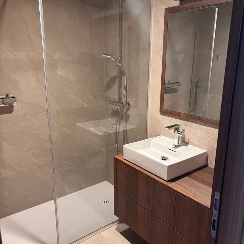 Das Bad ist hochwertig und modern ausgestattet und verfügt über elegante Steingutfliesen, ein Waschbecken von Laufen, maßgefertigte Tischlermöbel sowie eine großzügige Walk-in-Dusche. Die abgestimmten Materialien und die klare Gestaltung schaffen eine ruhige, hochwertige Atmosphäre.