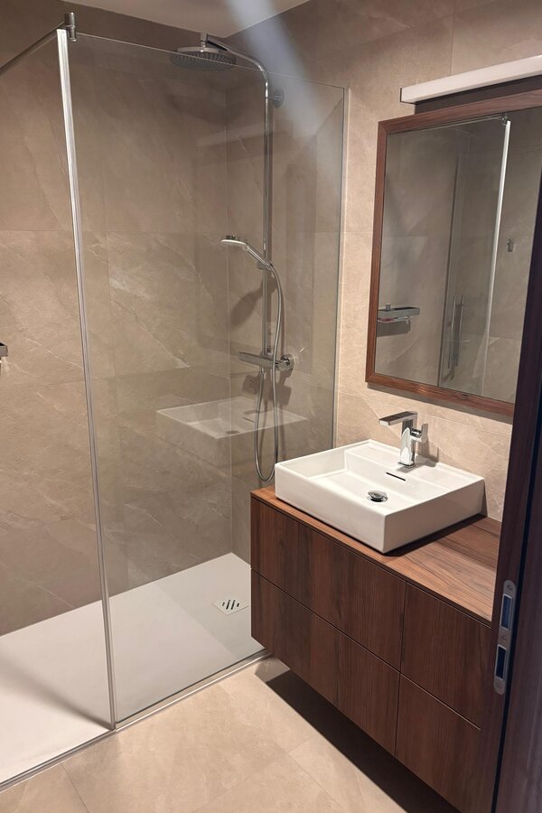 Das Bad ist hochwertig und modern ausgestattet und verfügt über elegante Steingutfliesen, ein Waschbecken von Laufen, maßgefertigte Tischlermöbel sowie eine großzügige Walk-in-Dusche. Die abgestimmten Materialien und die klare Gestaltung schaffen eine ruhige, hochwertige Atmosphäre.